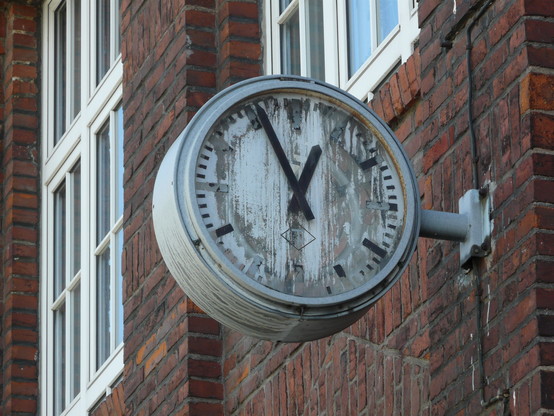 Eine alte Uhr mit Zifferblatt und Zeigern an einem alten Backsteingebäude, welche nicht mehr in Betrieb ist. Das Schutzglas fehlt bereits, das ehemals weise Zifferblatt ist bereits stark verwittert. Die Zeiger stehen dauerhaft auf zwölf Uhr und 56 Minuten.