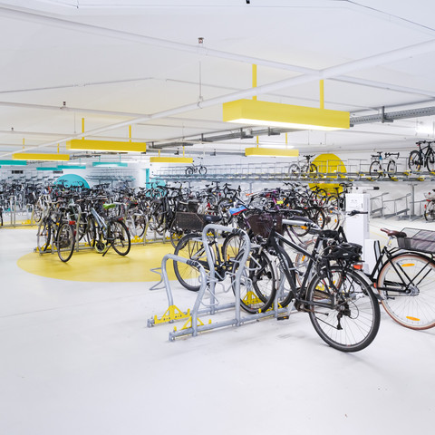 Ein Fahrradparkhaus in Karlsruhe. Boden und Wände sind aus hellem Beton mit gelben Farbakzenten. Es ist hell beleuchtet und sauber. Die Räder werden an stabilen Bügeln auf zwei Ebenen gelagert und angeschlossen.